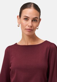 Top bordeaux a maniche lunghe con scollo a barca, texture a costine e cuciture delle spalle arricciate, abbinato a orecchini a cerchio dorati.