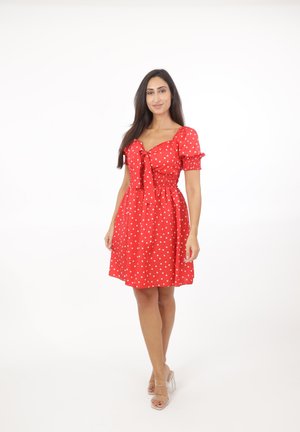 Donna in piedi che indossa un vestito rosso al ginocchio con pois bianchi, maniche a sbuffo, fiocco sul davanti e sandali con tacco trasparente su sfondo bianco.