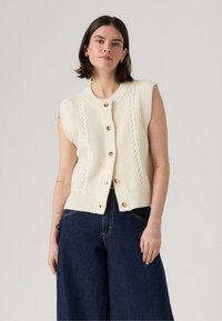 Cardigan sans manches crème avec une texture côtelée, un design en tricot torsadé et des boutons en bois. Assorti à un pantalon large en denim bleu foncé taille haute.