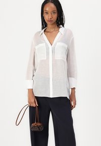 Camicia bianca in semitrasparente a maniche lunghe con due tasche sul petto e chiusura a bottoni, abbinata a pantaloni scuri a gamba larga e una borsa a pochette marrone.