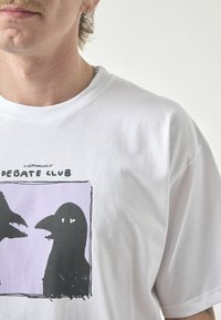 White cotton t-shirt featuring a graphic print of two black bird-like figures with eyes, set against a purple background.