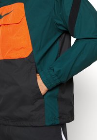 Persona vistiendo una chaqueta color bloque en verde, negro y naranja, con la mano en el bolsillo lateral, mostrando el puño elástico y el bolsillo frontal con solapa y logo.
