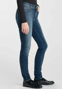 Jean skinny en denim bleu avec une légère décoloration, design à cinq poches et taille mi-haute, associés à des chaussures noires à enfiler.