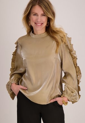 Beige satijnen blouse met gerimpelde mouwen, hoge hals en een ontspannen pasvorm. Gladde textuur met een subtiele glans, gecombineerd met donkere broek.