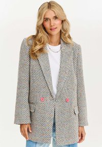 Blonďatá žena má na sobě multikolorový tweedový blejzr s růžovými knoflíky, bílou košili, korálkový náhrdelník a světle modré džíny na bílém pozadí.