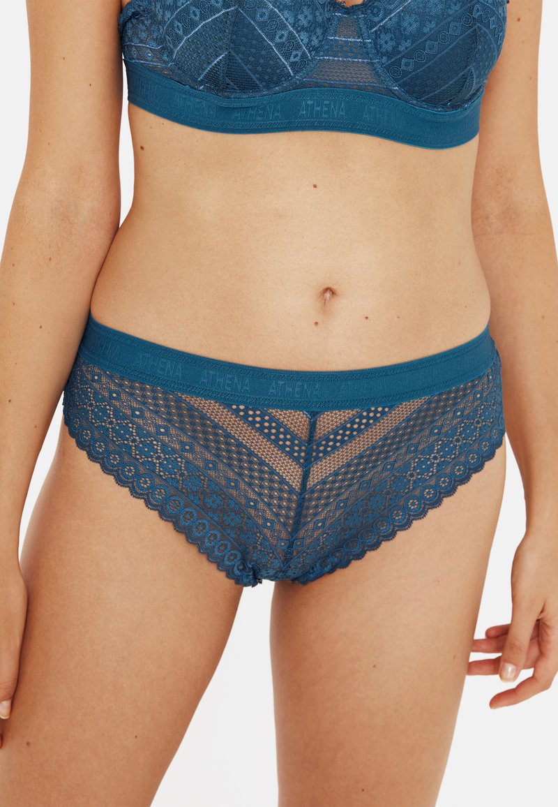 Des culottes en dentelle turquoise avec un ourlet festonné, un motif en chevrons et une large ceinture portant le logo "ATHENA". Texture délicate et semi-transparente.