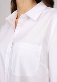 Chemise blanche à boutons avec un col, tissu texturé et une poche sur la poitrine gauche avec un logo brodé subtil.