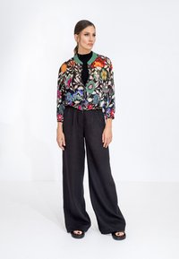 Giacca bomber ricamata con motivi floreali colorati, con colletto a coste verde, abbinata a pantaloni larghi in lino neri e sandali neri.