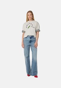 Witte korte mouwen shirt met zwarte opdruk en groene olijf grafiek, gecombineerd met high-waisted blauwe spijkerbroek en rode schoenen. Gestreepte mouwaccenten.