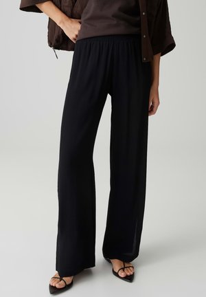 Vrouw draagt losse zwarte broek en zwarte sandalen met bandjes en een puntige neus, staand tegen een effen lichte achtergrond.