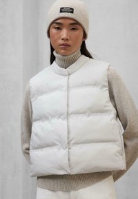 Chaleco acolchado blanco con forma cuadrada, que presenta un cuello alto y cierre de botones, superpuesto a un suéter de punto ribeteado en beige claro.