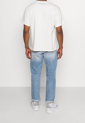 Camiseta de algodón blanca de manga corta, con el texto en la parte de atrás que dice "no es fácil ser verde", combinada con jeans de mezclilla azul claro y zapatillas.