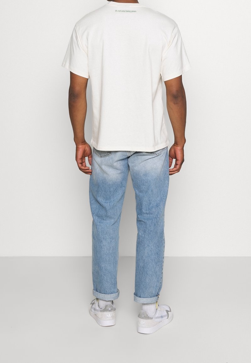 Vit bomull t-shirt med korta ärmar, text på ryggen som säger "det är inte lätt att vara grön", ihop med ljusblå denimjeans och sneakers.