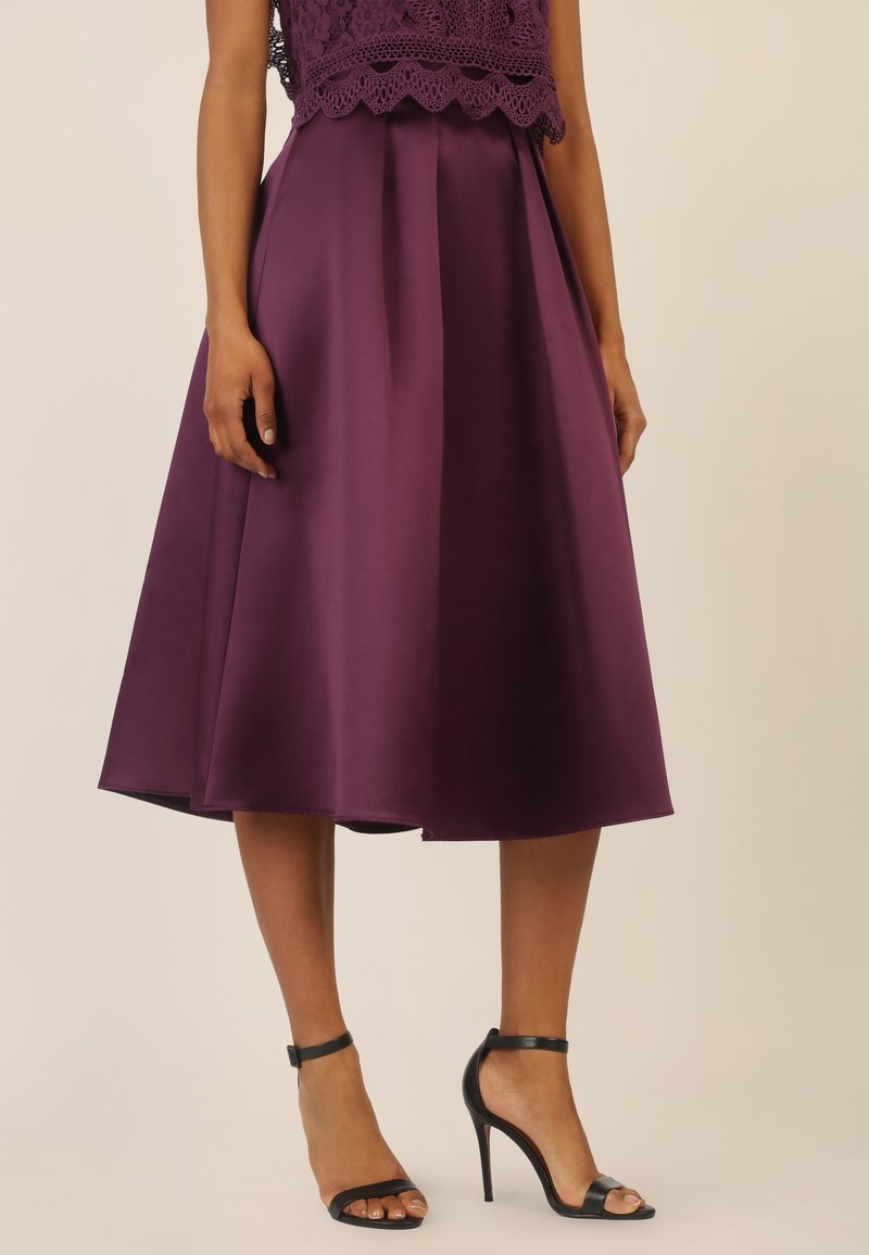 Gonna di raso viola con pieghe, lunghezza fino al ginocchio. Indossata con tacchi alti neri con cinturini. Presenta un top in pizzo, che aggiunge texture e dettagli.