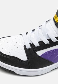 Hvit sneakers med svarte og lilla detaljer, laget av teksturert lær. Har perforeringer, hvite lisser og en polstret krage.