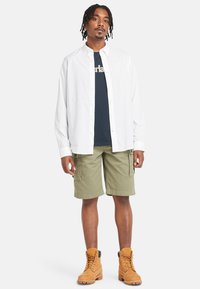 Witte shirt met knopen over een marineblauwe T-shirt, olijfgroene cargoshorts en tan kleurige veterschoenen. De outfit heeft een relaxte pasvorm en functionele zakken.