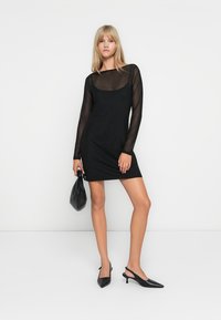 Rochie mini neagră cu mâneci lungi din material transparent, decolteu rotund, siluetă ajustată, asortată cu accesorii negre și pantofi cu vârf ascuțit.