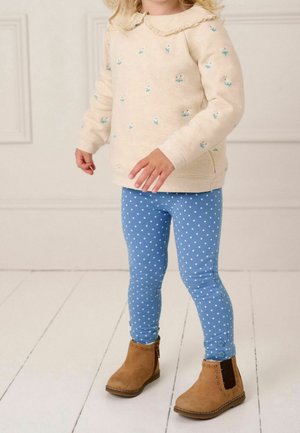 Enfant portant un sweat-shirt beige à fleurs, des leggings bleus à pois et des bottines marron, debout sur un sol en bois blanc.