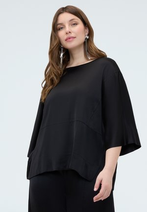 Kvinde med langt brunt hår iført oversized sort top med brede ærmer og store sølvfarvede hængende øreringe mod en ensfarvet baggrund