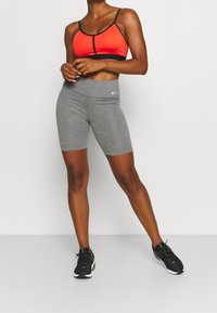 Brassière de sport rouge et noire associée à un short de cycliste taille haute gris, avec un logo Nike sur le short et des chaussures de sport noires.