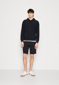 Marine hettegenser med frontlomme, kombinert med svarte shorts. Modell med hvite sneakers, minimalistisk design med myk tekstur.