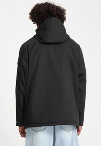 Personne aux cheveux bouclés portant une veste à capuche noire et un jean en denim bleu clair, vue de dos sur un fond blanc uni.