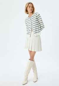 Sweter w paski w kolorze białym i szarym, zestawiony z plisowaną spódnicą w kolorze kremowym i wysokimi, błyszczącymi białymi butami do kolan. Prosty fason z guzikami.