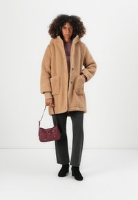 Beige, flauschiger, kapuzenartiger Mantel mit zwei Vorderaschen, kombiniert mit dunkelgrauen Hosen, schwarzen Stiefeletten und einer kastanienbraunen Handtasche.