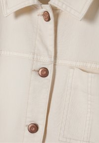 Krémová džínová bunda s měděnými knoflíky, viditelným šitím a náprsní kapsou. Texturovaná tkanina s hladkým povrchem.