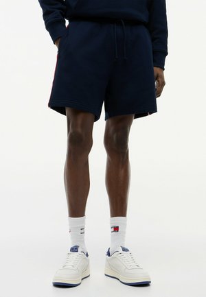 Ben af en person iført marineblå shorts med rød stribe, hvide sokker med logoer og hvide sneakers med blå detaljer på en hvid baggrund.