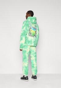 Πράσινο φούτερ με βάτες και κουκούλα, με γραφικό σχέδιο στην πλάτη, σε συνδυασμό με αντίστοιχο παντελόνι φόρμας, ολοκληρωμένο με μαύρα παπούτσια.