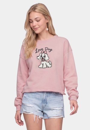 Kvinde med langt blondt hår iført en pink sweatshirt med en tegneseriehund og tekst "Love Dog," kombineret med lyseblå revne denimshorts.