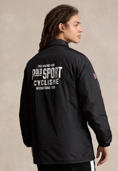 Schwarze Windbreaker-Jacke mit auffälligem weißen Text auf dem Rücken: "CYCLE-RACING-CLUB POLO SPORT CYCLISME INTERNATIONAL TOUR." Akzent mit britischer Flagge.