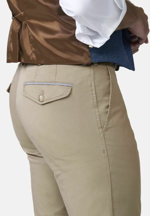 Mann, der beigefarbene Hose mit Klappentasche anpasst, kombiniert mit einer braunen Weste und sichtbarem weißen Hemdärmel vor weißem Hintergrund.