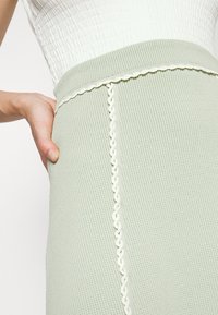 Personne portant une jupe texturée vert clair avec une bordure blanche et un haut blanc, main gauche posée sur la hanche, visible de la taille au milieu de la cuisse.