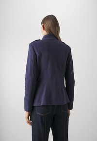 Chaqueta ajustada de color azul marino con cuello alto, diseño entallado, textura sutil y hombros estructurados. Detalles de costura visibles y puños.