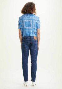 Camisa de manga corta azul con un estampado cuadrado, combinada con unos vaqueros oscuros, y presenta una etiqueta de cuero marrón claro en la cinturilla junto con zapatillas blancas.