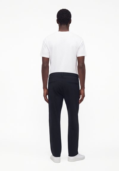 Homme vu de dos portant une chemise blanche unie à manches courtes, un pantalon foncé et des baskets blanches sur fond blanc.