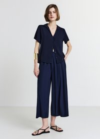 Chemise bleu marine à manches courtes avec des boutons, associée à un pantalon à jambes larges. Le modèle porte des sandales noires et un bracelet manchette doré.