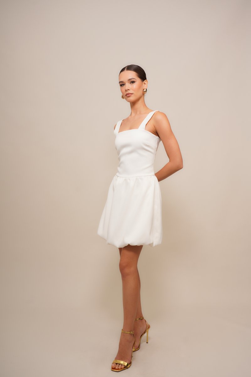White dress with a fitted bodice, square neckline, and flared, pleated skirt. Complemented by golden strappy sandals. Smooth texture.