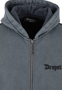 Sweat à capuche gris foncé de taille moyenne avec fermeture éclair complète et étiquette de marque "Dropsize" sur le curseur de la fermeture éclair ainsi qu'une broderie sur la poitrine.