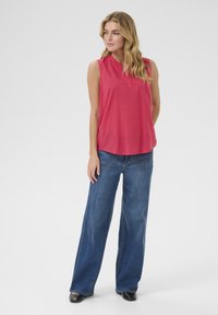 Ermeløs, lyserød bluse med en subtil tekstur og V-udskæring, parret med brede blå jeans og sorte sko.