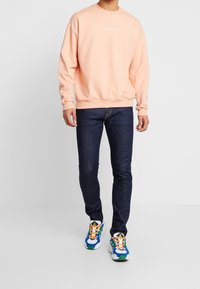 Man som bär en persikofärgad sweatshirt med vit "KAOTIKO" text, mörkblå jeans och färgglada blå, gröna och orange sneakers som går framåt.