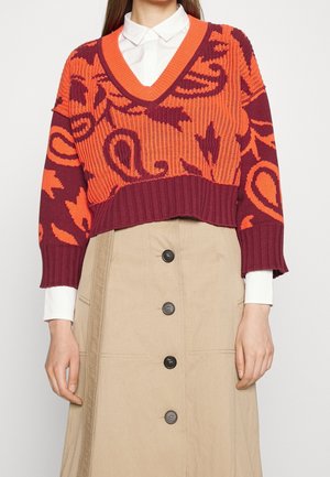 Frau trägt ein weißes Hemd mit Kragen unter einem orangen und bordeauxfarben gemusterten kurzen Pullover mit einem beigen Rock mit Knöpfen.
