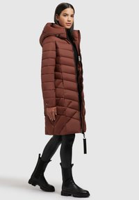 Eine Frau trägt einen langen braunen gesteppte Mantel mit Kapuze, schwarze Leggings und schwarze klobige Stiefel und steht vor einem schlichten hellen Hintergrund.