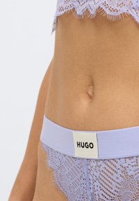 Lila čipkasto spodnje perilo z valovitim robom in gladkim pasom z belo etiketo, na kateri je napis "HUGO" v črni pisavi.