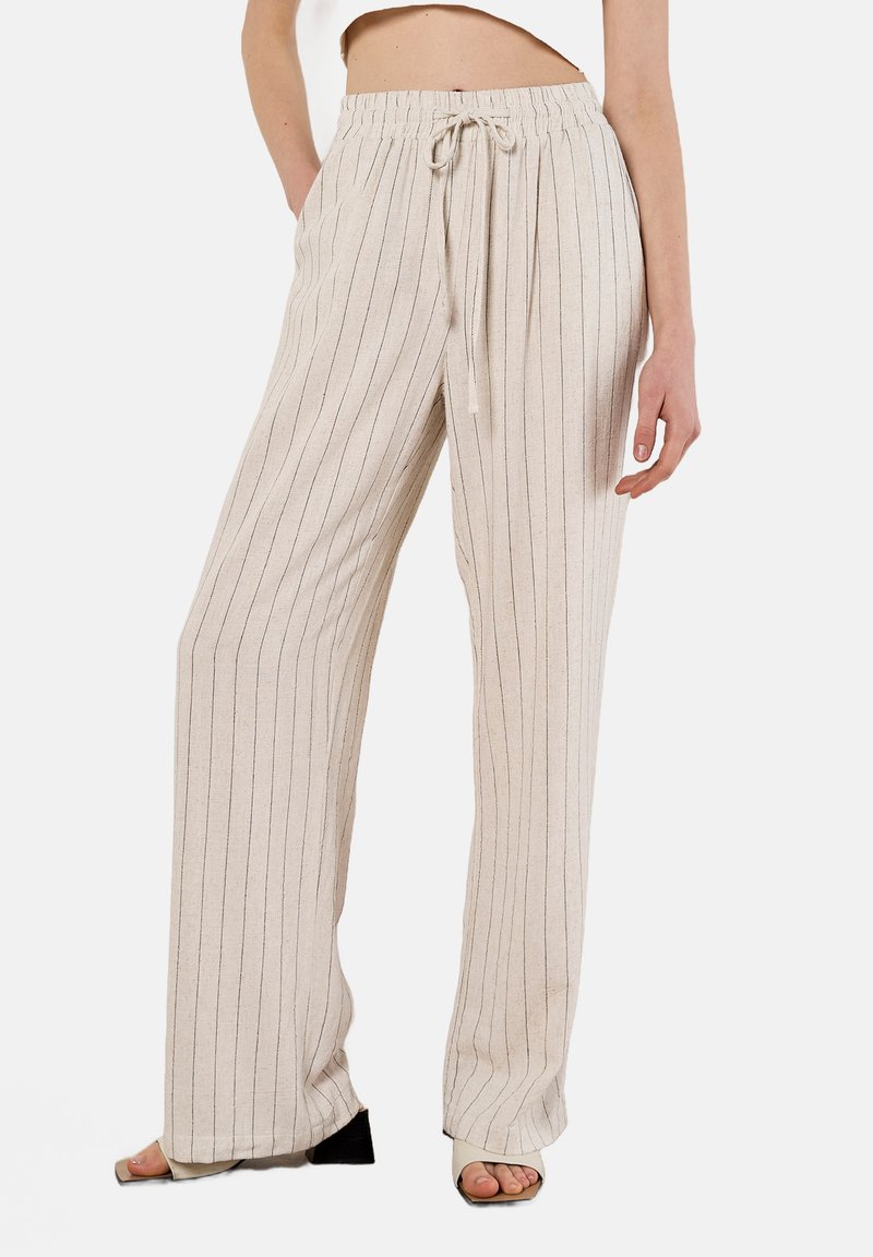Pantalons larges beige clair en tissu texturé avec des rayures verticales sombres. Dotés d'une taille élastique avec un cordon de serrage.