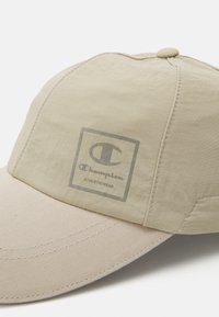 Beige caps laget av glatt stoff med en buet skygge. Har en grå Champion-logo foran. Inkluderer synlige sømmer og en toppknapp.