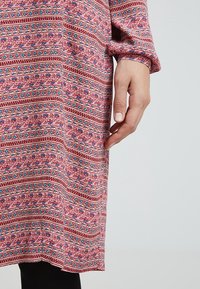 Une robe rose à motifs floraux et géométriques, avec des manches longues et un tissu texturé. Les détails incluent des rayures horizontales.