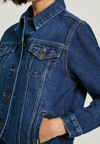 Giacca di jeans in blu scuro con colletto a punta, chiusura con bottoni, due tasche sul petto e cuciture a contrasto. Presenta una vestibilità comoda e maniche lunghe.